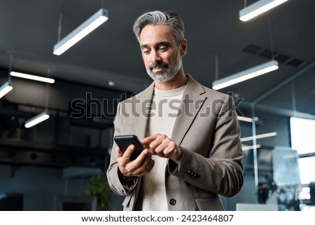 Busy mature older business man wearing suit using mobile cell phone at work. Middle aged businessman executive manager or entrepreneur holding smartphone looking at cellphone standing in office. Royalty-Free Stock Photo #2423464807