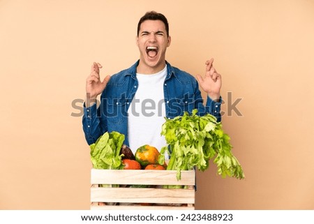 Farmer with freshly picked vegetables in a box isolated on beige background with fingers crossing