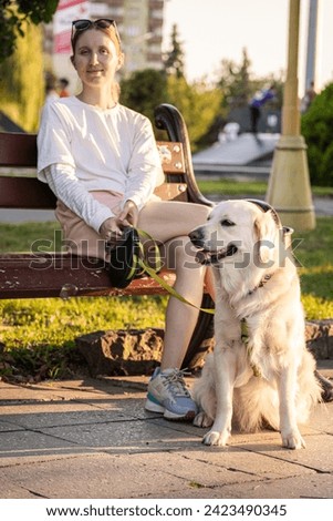 The dog owner and the golden retriever obediently sit on a bench in the park and enjoy a sunny, summer day. Royalty-Free Stock Photo #2423490345