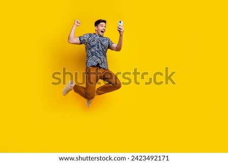 Full size photo of overjoyed guy wear stylish shirt brown pants look at smartphine jumping win lottery isolated on yellow color background