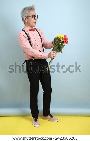 Asian senior man in black suspenders with red bow with gesture of holding a bouquet of flowers isolated on blue background. St Valentine's Day, Women's Day, Birthday