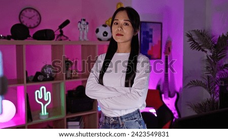 A young asian woman stands confidently with folded arms in a neon-lit gaming room, emanating a serene but focused demeanor. Royalty-Free Stock Photo #2423549831