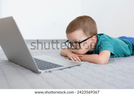 The child, leaning on his hands, look at the screen of the laptop. The child is watching cartoons, films or educational programs with concentration. High quality photo