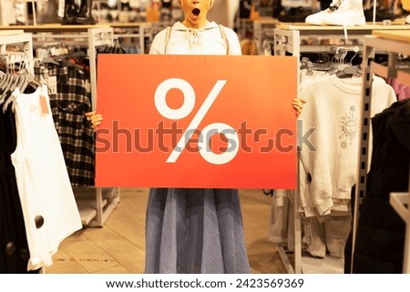 A girl in a clothing store holds a large red banner with interest in her hands. A woman has emotions of surprise and joy on her face. Discounts in the store, promotions, sale, Black Friday.