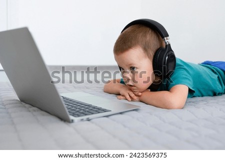 A little boy in headphones is lying down watching cartoons or educational programs in a laptop. Education, laptop, headphones, learning new things, entertainment. High quality photo