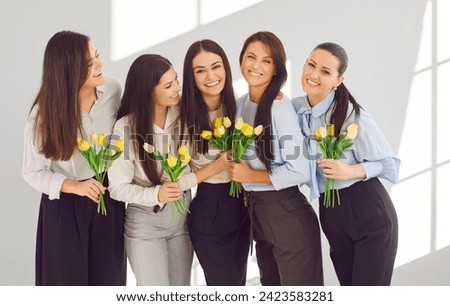 Group of happy business women in office celebrating International Women's Day together. Portrait of stylish and beautiful businesswomen with yellow tulips hugging and smiling in front of camera. Royalty-Free Stock Photo #2423583281
