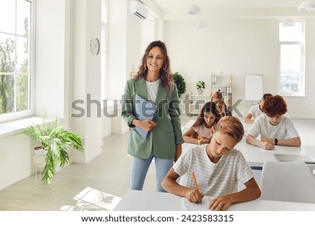 Young happy woman teacher standing in a school classroom holding book in hand. Her smiling face creating an inviting atmosphere for education and showing essence of a dynamic lesson. Royalty-Free Stock Photo #2423583315