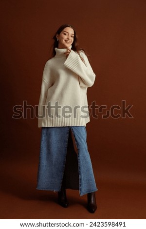 Fashionable happy smiling woman wearing oversized white turtleneck sweater, maxi blue denim skirt, pointed toe ankle leather boots, posing on brown background. Full-length studio fashion portrait Royalty-Free Stock Photo #2423598491