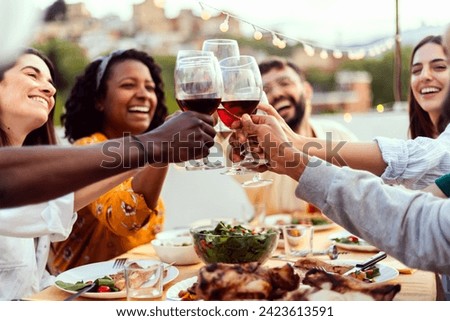 Young group of people enjoying dinner reunion in the evening at rooftop home. Millennial diverse friends social gathering reunited on terrace table, having fun celebrating barbecue party at night.