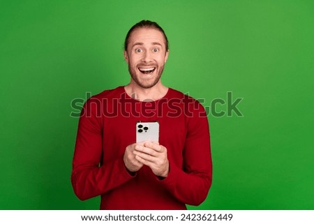 Portrait of satisfied impressed guy with long hair wear red shirt hold smartphone asstonished staring isolated on green color background