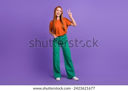 Full length photo friendly and peaceful ginger lady in orange t shirt green pants makes v sign to you isolated on purple color background