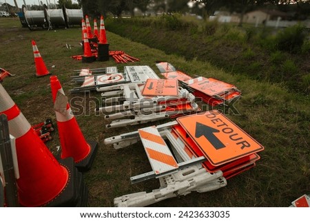 Multiple orange and black detour ahead signs barricades and orange cones in a  grass field. Also has black and white signs and three large rolls of electrical wire in the background in sun.