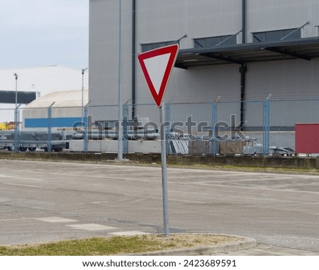 Yield (give way priority) road sign attached on a metal pole with a street and industrial building in the background
