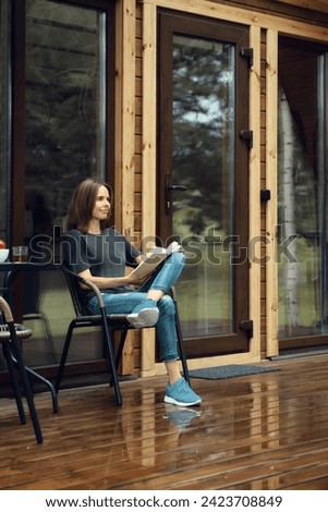 Cute woman in casual clothing sits reading on a wet terrace of a log cabin after the rain, enjoying fresh air of woods Royalty-Free Stock Photo #2423708849