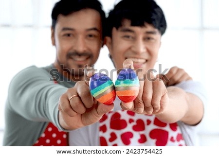 Happy young LGBT couple holding rainbow Easter eggs that are symbol of LGBTQ flag color to camera, Asian gay male lover has special moment together to celebrate Easter holiday. Sweet lover has dating.