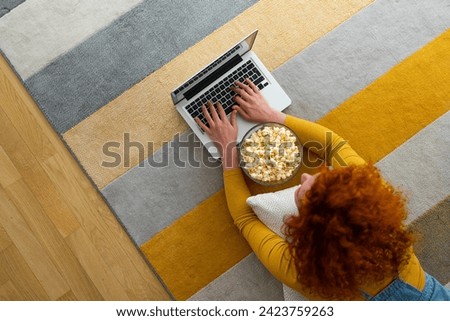Photo of female person taken from above. Woman with red curly hair lying on the carpet and typing on laptop in cozy home atmosphere.