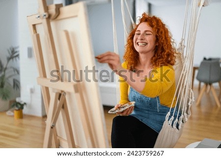 Young art school student sitting in a atelier, holding paint palette and painting on empty canvas while smiling. Royalty-Free Stock Photo #2423759267