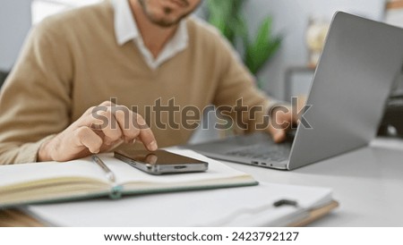 Young hispanic man multitasking with smartphone and laptop in a modern office setting.