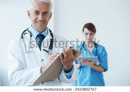 Portrait, mature doctor writing on his clipboard and with a nurse browsing on a digital tablet in the background. Senior doctor and nurse standing in corridor looking at files discussing reports