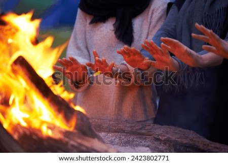 Hands, fire and people camping outdoor, keeping warm with open flame at campsite, friends huddle for heat and bonding. Travel, nature and relax around campfire, community and together for adventure Royalty-Free Stock Photo #2423802771