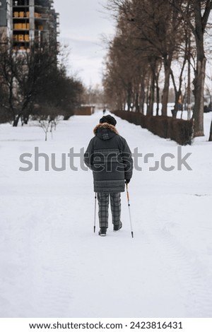 An adult, elderly woman with aluminum hiking poles walks in the winter on white snow on the street outdoors. Photography, healthy lifestyle concept.