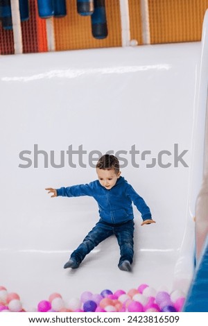 Happy boy, a small child goes down, slides down while sitting on a plastic slide in the playroom into a pile of colored balls. Photography, childhood concept.