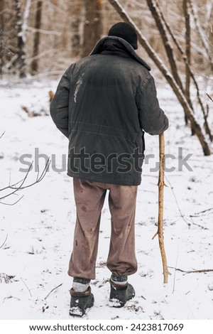 An old, elderly lonely forester, a homeless beggar man in dirty clothes walks through the forest with a wooden stick in search of food, shelter in the cold snowy winter. Photography, poverty concept.
