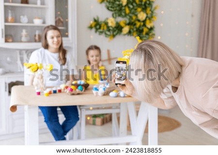 Mom takes pictures of two daughters sitting at kitchen at home during Easter holidays. Easter preparation coloring eggs together taking selfie photo on smartphone smiling cheerful