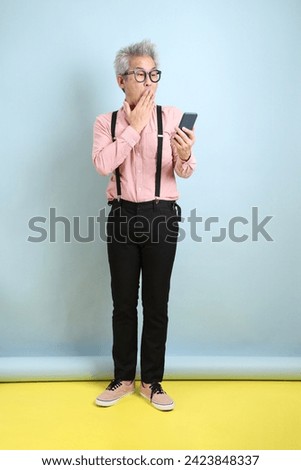 Asian senior man in black suspenders with red bow with gesture of Holding mobile phone isolated on blue background. St Valentine's Day