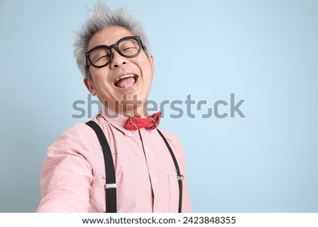 Asian senior man in black suspenders with red bow with gesture of taking a selfie isolated on blue background. St Valentine's Day