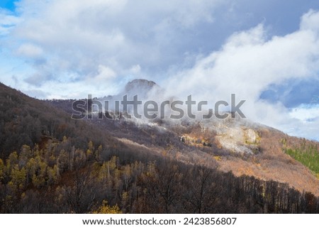 Beautiful autumn colorful view of a mountain rocky peak surrounded by a white cloud and a forest with dried leaves - aerial drone photo