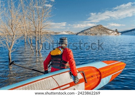 male paddler wearing a drysuit, life jacket and safety leash  is starting workout on a long racing stand up paddleboard on a mountain lake in Colorado - Horsetooth Reservoir in winter conditions Royalty-Free Stock Photo #2423862269