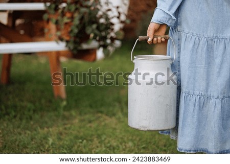 A caucasian woman in a blue dress holds an aluminum milk can in a lush garden. Concept for organic farming, fresh dairy products, or rustic lifestyle. Copy space available. Royalty-Free Stock Photo #2423883469
