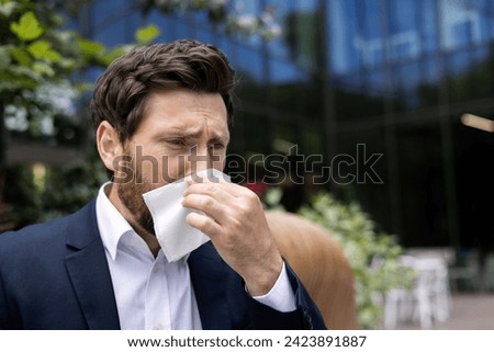 Close-up photo of a sick male businessman standing outside an office center, wearing a suit, wiping his nose with a napkin, suffering from a runny nose and a cold.