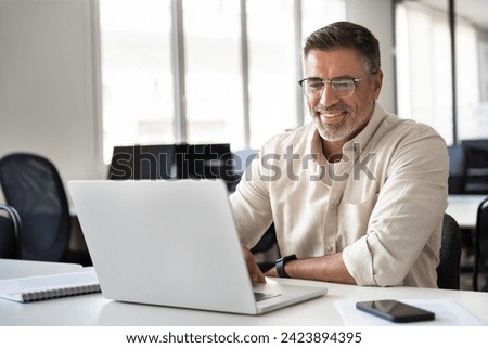 Happy smiling mature Indian or Latin business man ceo trader using computer, typing, working in modern office, doing online data market analysis, thinking planning tech strategy looking at laptop. Royalty-Free Stock Photo #2423894395