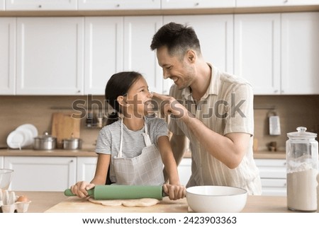 Cheerful baker father touching face of adorable daughter girl in apron with floury finger, having fun, laughing, rolling dough on home kitchen table, baking homemade bakery food Royalty-Free Stock Photo #2423903363