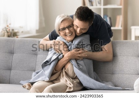 Positive young adult son man caring for happy elderly mature mother sitting on comfortable sofa, throwing warm plaid, scarf on moms shoulders, hugging from behind with love, affection Royalty-Free Stock Photo #2423903365