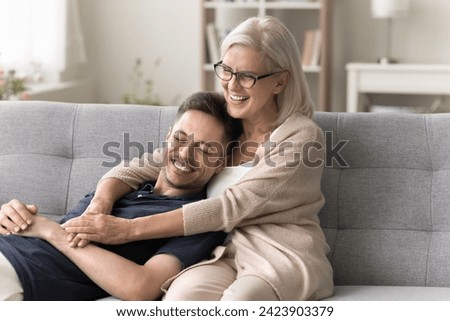 Joyful laughing senior mom hugging cheerful handsome son with care, resting on sofa. Happy mother and adult child meeting at home, having fun, enjoying family leisure, comfort Royalty-Free Stock Photo #2423903379