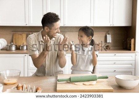 Joyful preteen kid girl and happy dad having fun while baking in home kitchen, clapping floury hands, making white powder cloud over table with bakery ingredients, laughing Royalty-Free Stock Photo #2423903381