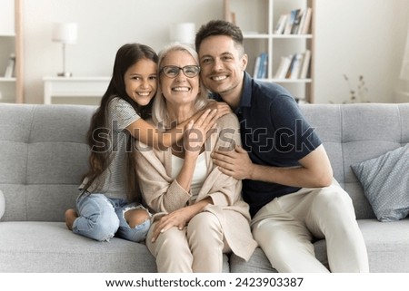 Happy sweet granddaughter kid and young dad hugging beloved grandma. Cheerful preteen girl, father and grandmother posing for family picture on home couch, looking at camera, smiling
