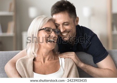 Happy son man hugging positive excited mature mom from behind, embracing mum in glasses with love, gratitude, joy, talking, laughing, having fun. Mother and adult child casual home portrait