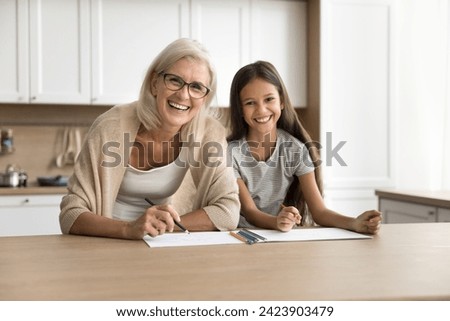 Happy pretty blonde grandma and adorable preteen grandkid girl drawing together at home, sitting at table with paper and colorful pencils, looking at camera, laughing , smiling for portrait