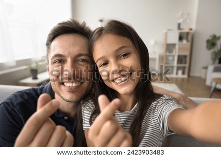 Happy young handsome dad and adorable daughter kid showing finger hearts, looking at camera with toothy smiles, taking selfie picture at home with symbols of peace, charity, love