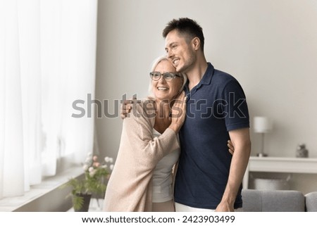 Cheerful dreamy adult son and senior mom standing close at window, hugging with love, care, looking away, smiling, talking, discussing leisure, family plans, retirement. Home casual portrait Royalty-Free Stock Photo #2423903499