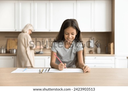 Happy preteen kid girl drawing in colorful pencils in home kitchen while blonde grandma cooking lunch in blurred background. Positive schoolchild doing school homework task at home