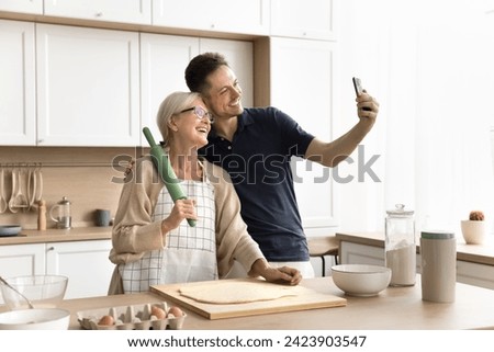 Happy senior mother and adult son taking cooking selfie on mobile phone, baking pastry dessert in home kitchen, posing at table with bakery ingredients, having fun, using internet technology