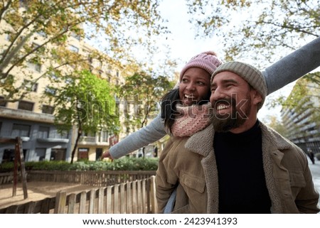 Portrait of beautiful multiracial happy couple outdoor young woman and man in love hugging and smiling in piggyback pointing somewhere at street in christmas time. People dressed in winter clothes