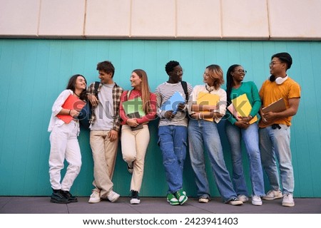 Happy international students posing outdoors. Smiling looking each other. Young people in multicultural community of friends and embracing with backpacks and workbooks in a blue wall background
