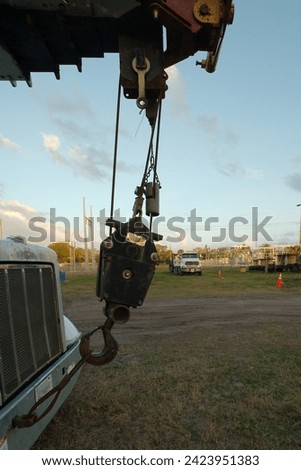 Close up vertical of black Hook Block from a white crane truck. In a grass field with other truck and green trees in the background. Has a bright sky and room for copy. Royalty-Free Stock Photo #2423951383
