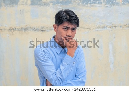 An Asian man is contemplating wearing a sky-blue shirt against the backdrop of a deteriorating yellow wall.